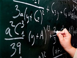 An image of a chalkboard with math equations written all over it and a person's hand writing on it.