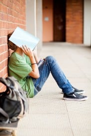 An image of a kid sitting against a wall, while holding a notebook over his face.